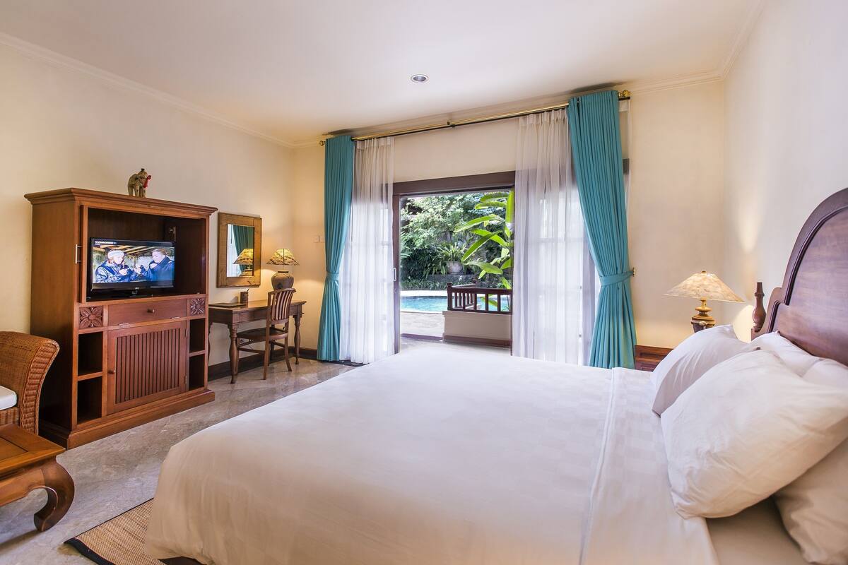 A hotel room with a large bed, a desk with a chair, a television, and a view of a pool and greenery.
