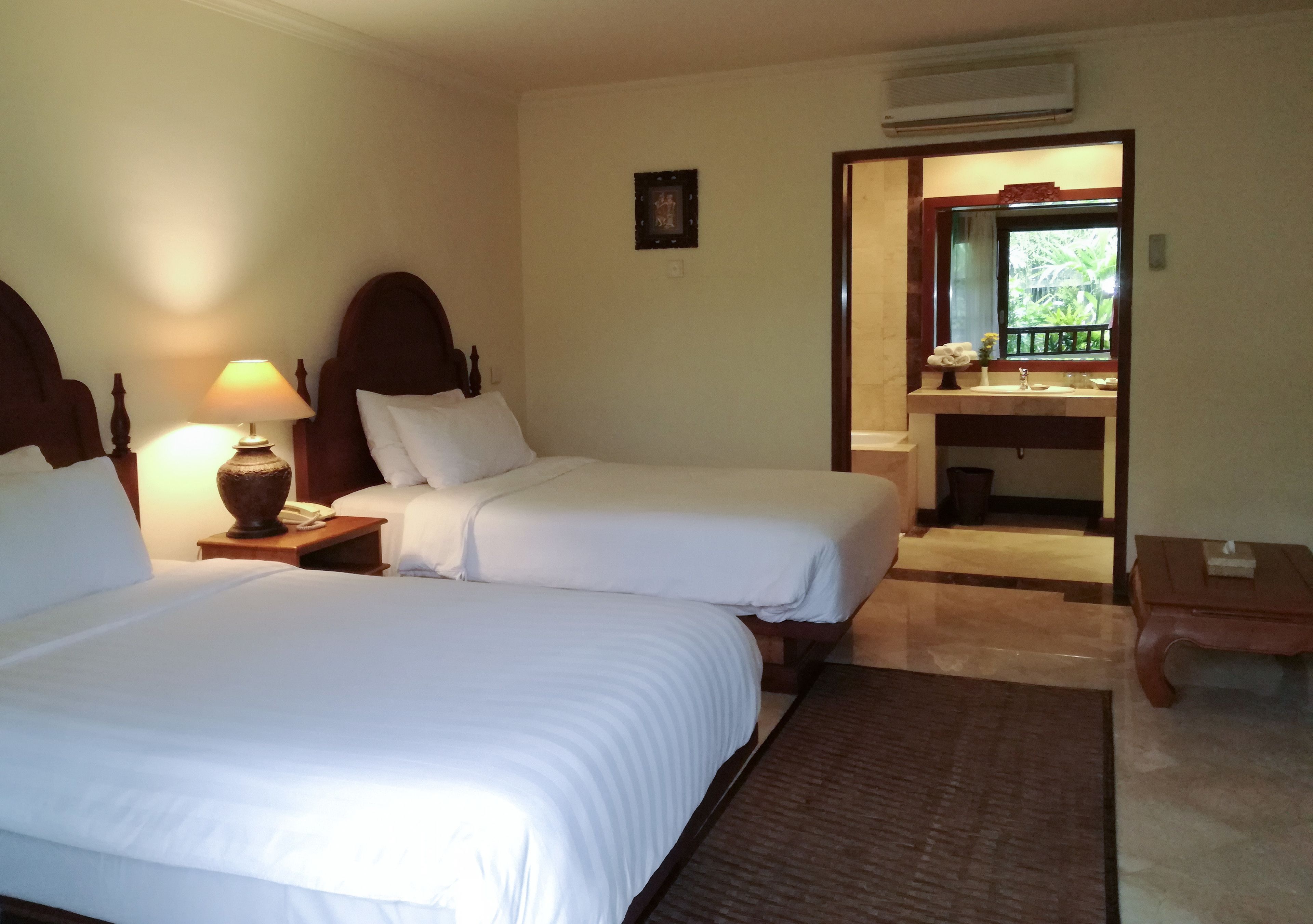 Habitación de hotel con dos camas, una mesita de noche con lámpara, un baño con espejo y una pequeña mesa con un florero.