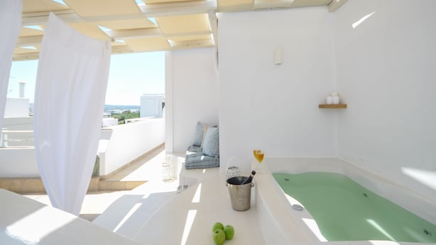 A bright, white room with a green bathtub, a small table with a bucket and apples, and a view of the city. - Blue Sky Summer (Naxos)