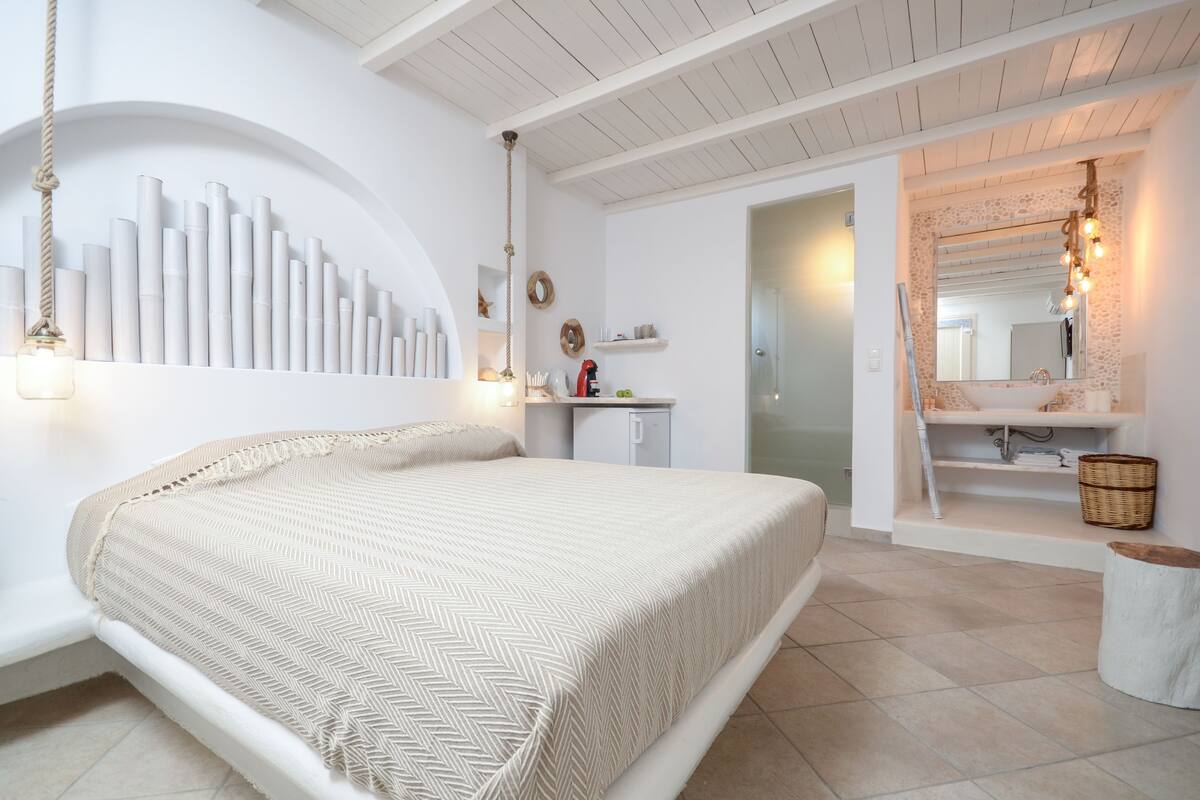 A bedroom with a large bed, a white wardrobe, a bathroom with a sink and mirror, and a hanging light fixture.