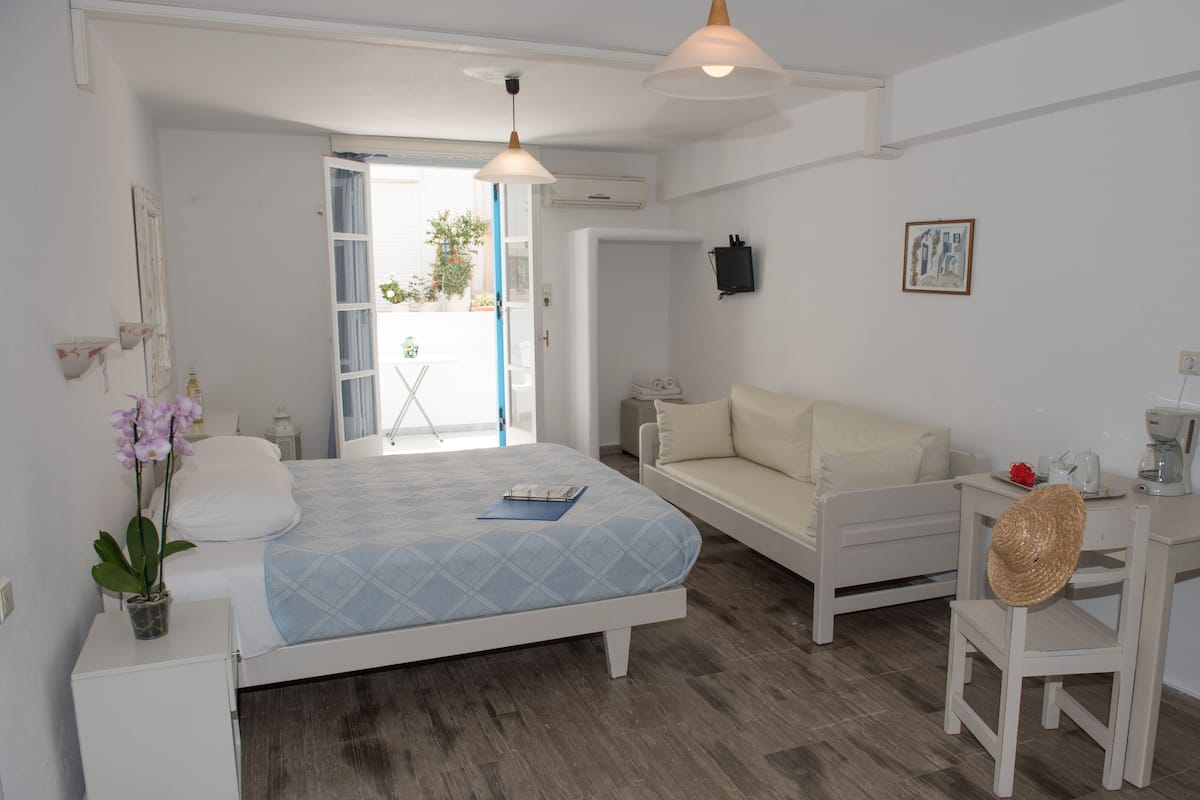 A bedroom with a bed, a sofa, a desk, and a chair. There is a window, a door, and a wall-mounted picture.