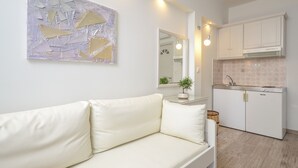 A modern living room with a white sofa, a minimalist abstract painting, and a kitchen with white cabinets and a tiled backsplash. - Blue Sky Summer (Naxos)