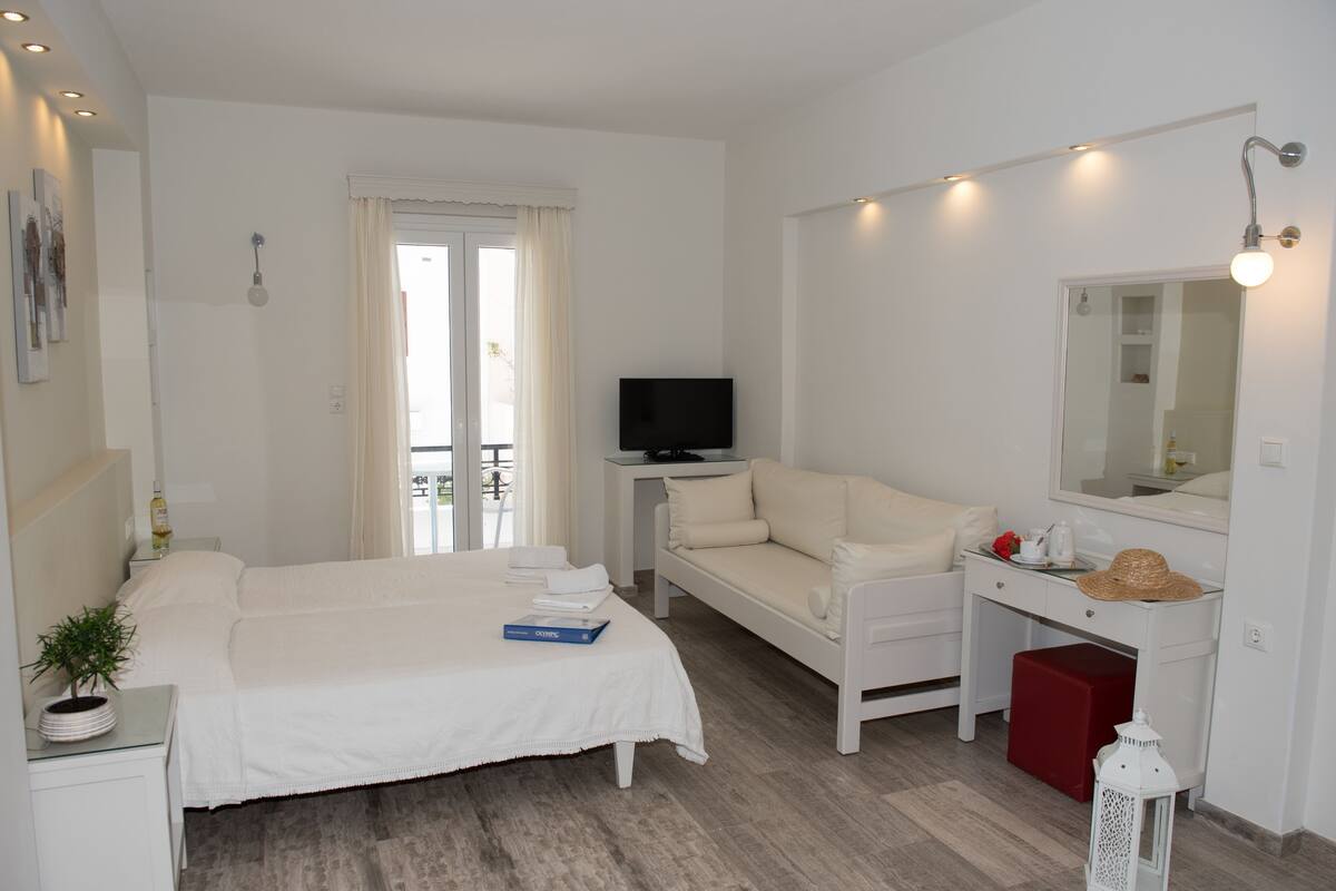 A hotel room with a bed, a sofa, a desk with a TV, a mirror, and a window with curtains.