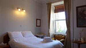Una camera d'albergo con un letto rifatto, un comodino con una lampada, un piccolo tavolino con un vassoio di oggetti e una finestra con tende.