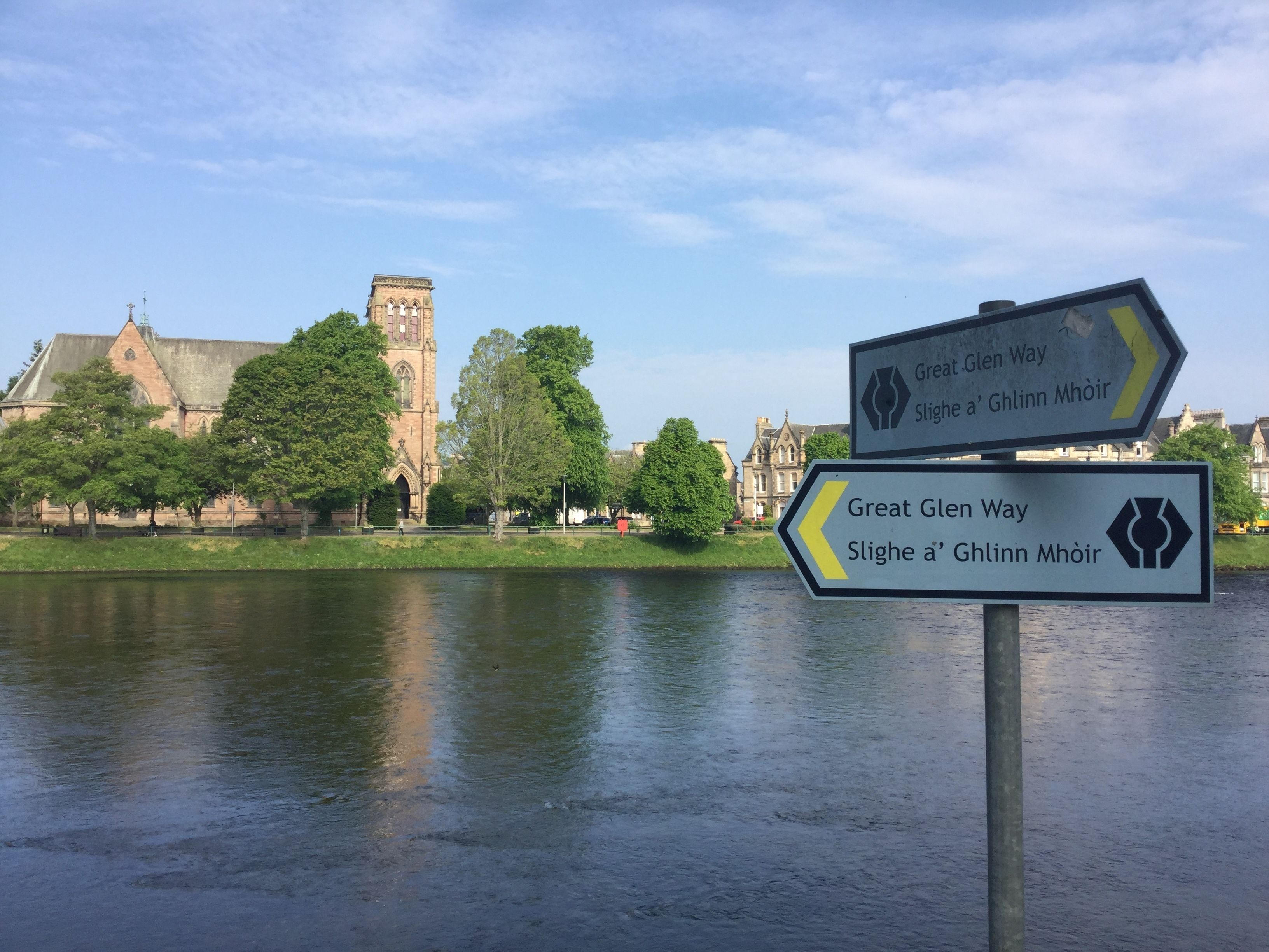 A signpost with directions to Great Glen Way and Slighe a' Ghlinn Mhóir, with a river and historic buildings in the background.