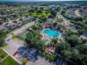 Ett flygbildsbild av ett bostadsområde med en swimmingpool omgiven av hus, en tennisbana och en parkeringsplats.