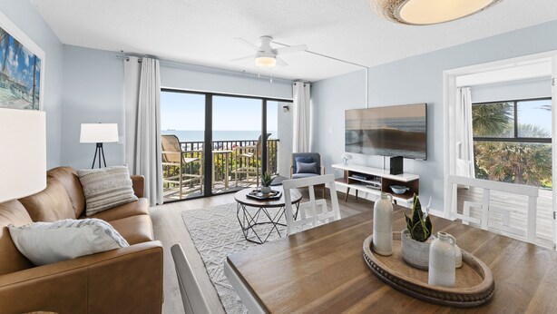 A modern living room with a sofa, TV, and dining area. - Coastal Escape Oceanfront Corner Unit with Heated Pool and Resort Amenities (Cocoa Beach)