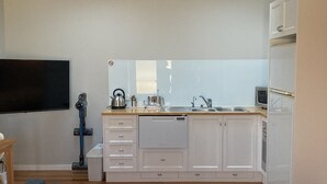 A modern kitchen with white cabinets, a built-in oven, and a sink. There is a flat-screen TV mounted on the wall, and a vacuum cleaner is visible. - Bella Loft:  A studio apartment in the heart of Woodend. (Woodend)