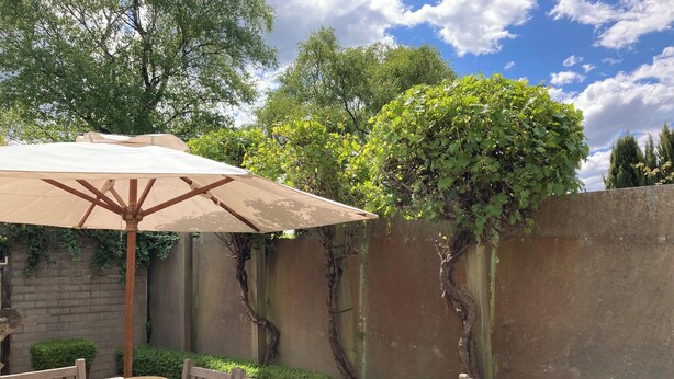 An outdoor patio with a wooden table and chairs, a beige umbrella, and a brick wall with climbing plants. - Bella Loft:  A studio apartment in the heart of Woodend. (Woodend)