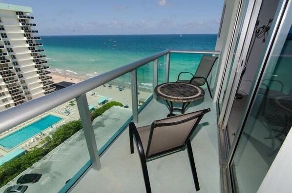 Un balcon doté d’une balustrade en verre, une table ronde et deux chaises. Le balcon offre une vue sur une piscine et une plage aux eaux d’un bleu limpide.