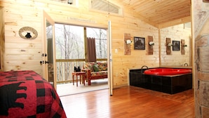 A wooden cabin interior with a bed, a sofa, a red bathtub, and a balcony with a view of trees.