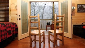 A cozy cabin interior with a wooden floor, a bed with a red and black checkered blanket, and a view of the snowy forest through a glass door.