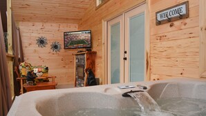 A wooden cabin interior with a hot tub, a welcome sign, and a television displaying a sports game.