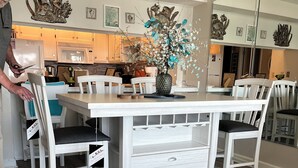 A dining area with a white table and chairs, a vase with flowers, and a kitchen in the background. - INCREDIBLE OCEANFRONT VIEWS/3 BR, 3 Bath Luxury Condo/Sea Marsh I (North Myrtle Beach)