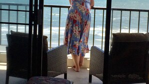 A woman in a floral dress standing on a balcony with ocean views, two wicker chairs, and a small table. - INCREDIBLE OCEANFRONT VIEWS/3 BR, 3 Bath Luxury Condo/Sea Marsh I (North Myrtle Beach)