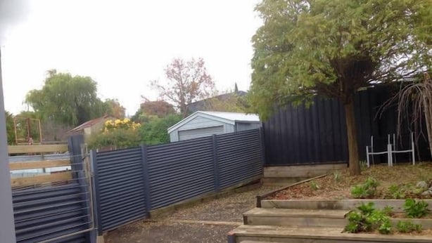 Een achtertuin met een houten terras, een bloembed en een metalen hek.