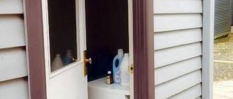 A small, enclosed outdoor area with a door leading to a white interior space, possibly a bathroom, with a white countertop and a bottle of liquid.