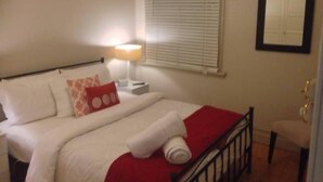 Een net opgemaakt bed met witte lakens en een rode kussen, een bedlampje en een raam met jaloezieën.
