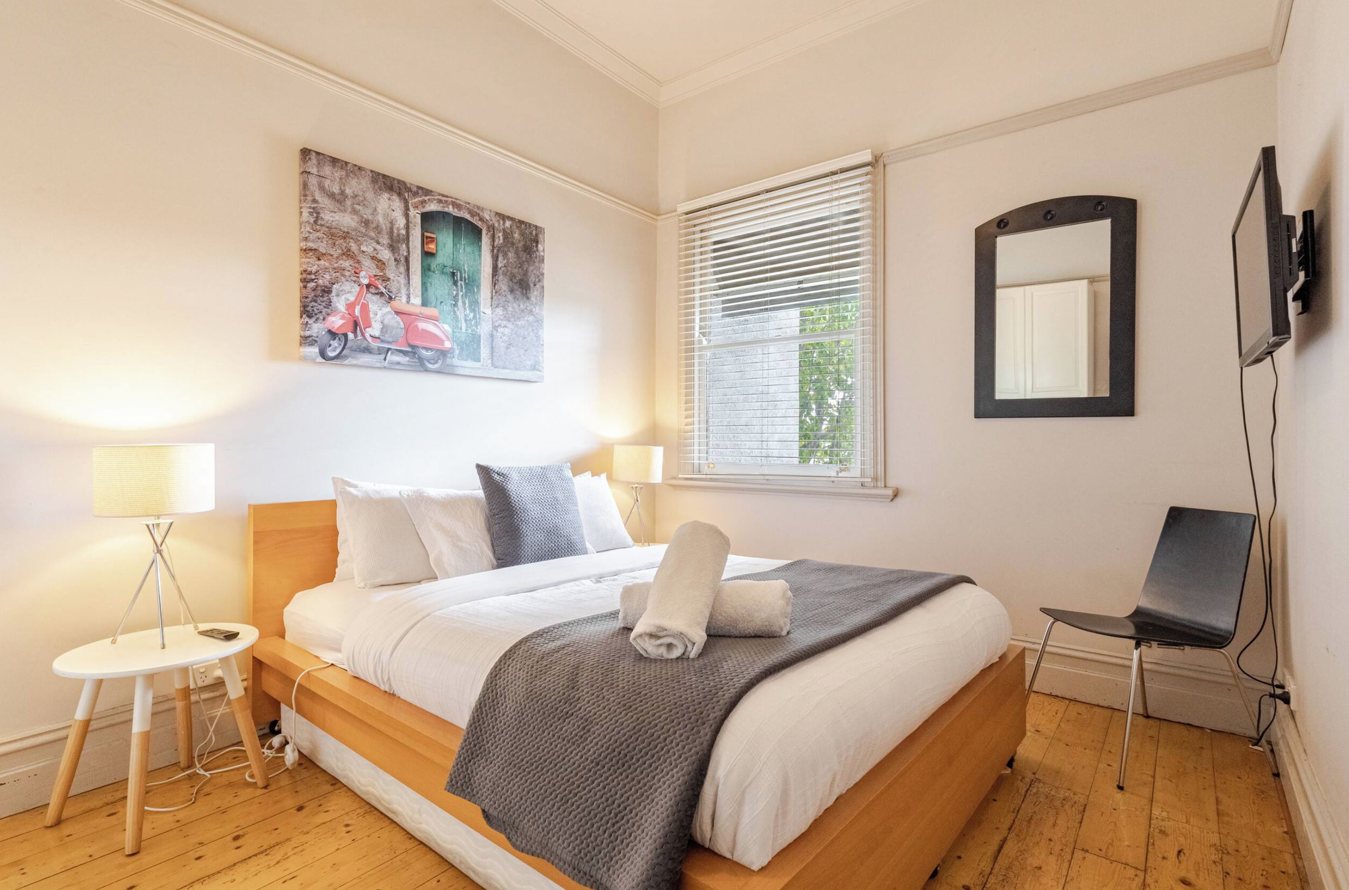 A neatly arranged bedroom with a bed, bedside lamps, a chair, a window with blinds, and a wall-mounted TV.
