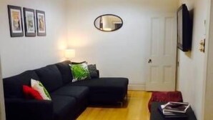 Een woonkamer met een zwarte modulaire bank, een klein zwart tafeltje met boeken, een ronde spiegel aan de muur en een houten vloer.