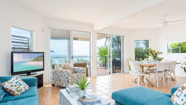 A spacious living area with a large TV, blue sofas, a dining table with chairs, and a balcony with a view of the ocean. - Moreton Bay Beach LodgeUnwind in  indulge in discovery (Cleveland)