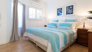 A bedroom with a bed, curtains, a window, an air conditioner, a nightstand, a lamp, and two framed pictures on the wall. - Moreton Bay Beach LodgeUnwind in  indulge in discovery (Cleveland)
