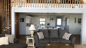 Una sala de estar moderna con un sofá gris, una mesa de centro y un comedor con una mesa azul. Se ve una cocina al fondo con armarios blancos y un letrero que dice "BEACH HOUSE".
