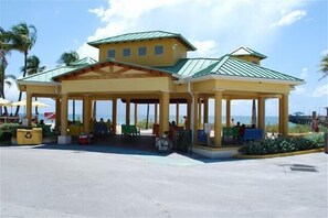 A beachside pavilion with a green roof and yellow walls, featuring outdoor seating and a bike rack.