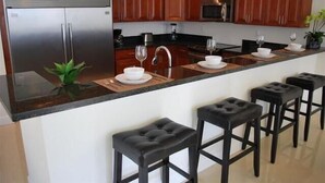 A modern kitchen with wooden cabinets, a stainless steel refrigerator, a microwave, and a bar area with stools.