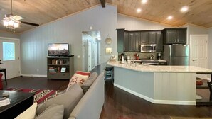 A spacious living room with a large sofa, a television, and a bookshelf. A kitchen area with dark cabinets and a stainless steel refrigerator is visible. - Just the Right Size. Exactly Enough | Highlands Haven (Highlands)