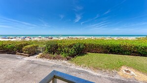 Vista sulla spiaggia con cielo sereno, una zona verde con recinzione metallica e un vialetto lastricato che conduce alla spiaggia.