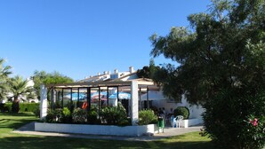 A glass-enclosed outdoor seating area with umbrellas, surrounded by greenery and a white building in the background.