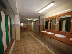 Ein langer Gang mit mehreren Toilettenkabinen, einem zentralen Waschbereich mit Spiegeln und einem gefliesten Boden.
