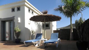 Eine Terrasse mit zwei Liegestühlen, einem Reedschirm und Topfpflanzen.