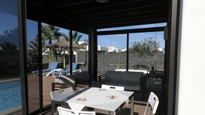 Ein überdachter Außenbereich mit Blick auf einen Pool, weiße Stühle und ein Tisch mit einer blumigen Tischdecke.