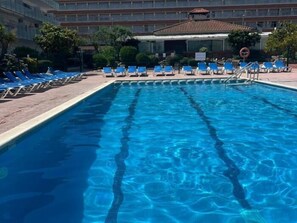 A rectangular swimming pool with clear blue water, surrounded by blue lounge chairs and a brick walkway. - APCOSTAS Lotus / One Bedroom (Blanes)