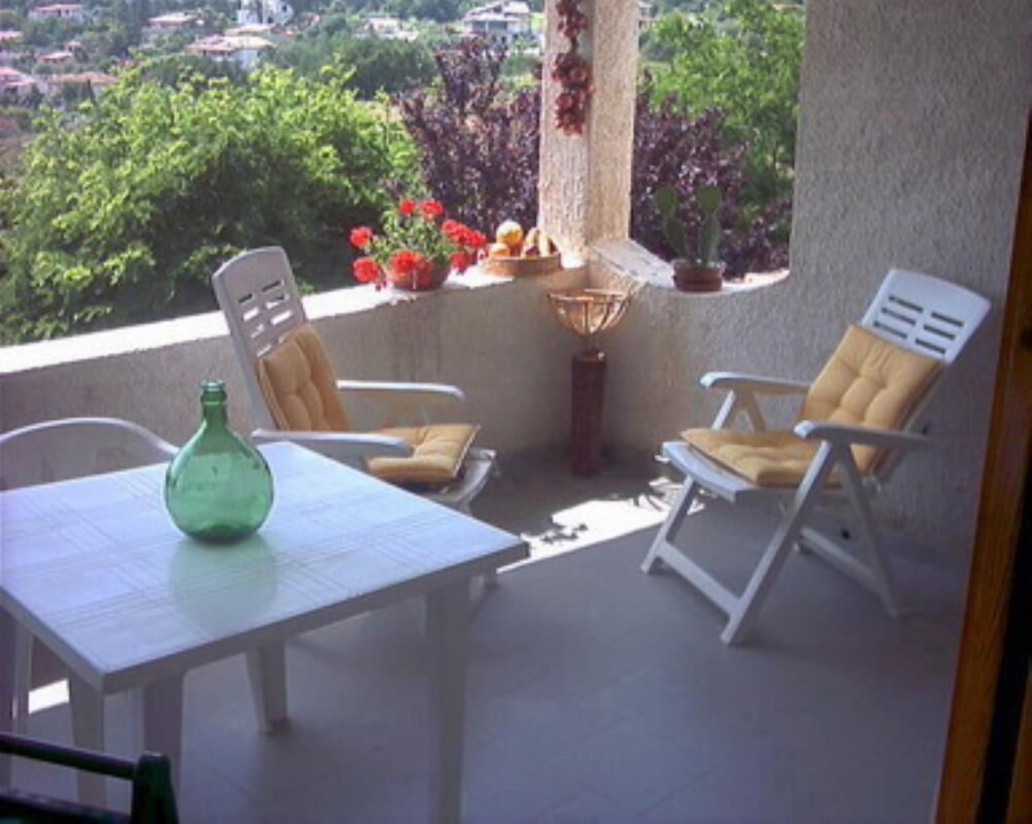 A balcony with two white chairs, a small table, and a green bottle. There are potted plants and a view of greenery and buildings in the background.