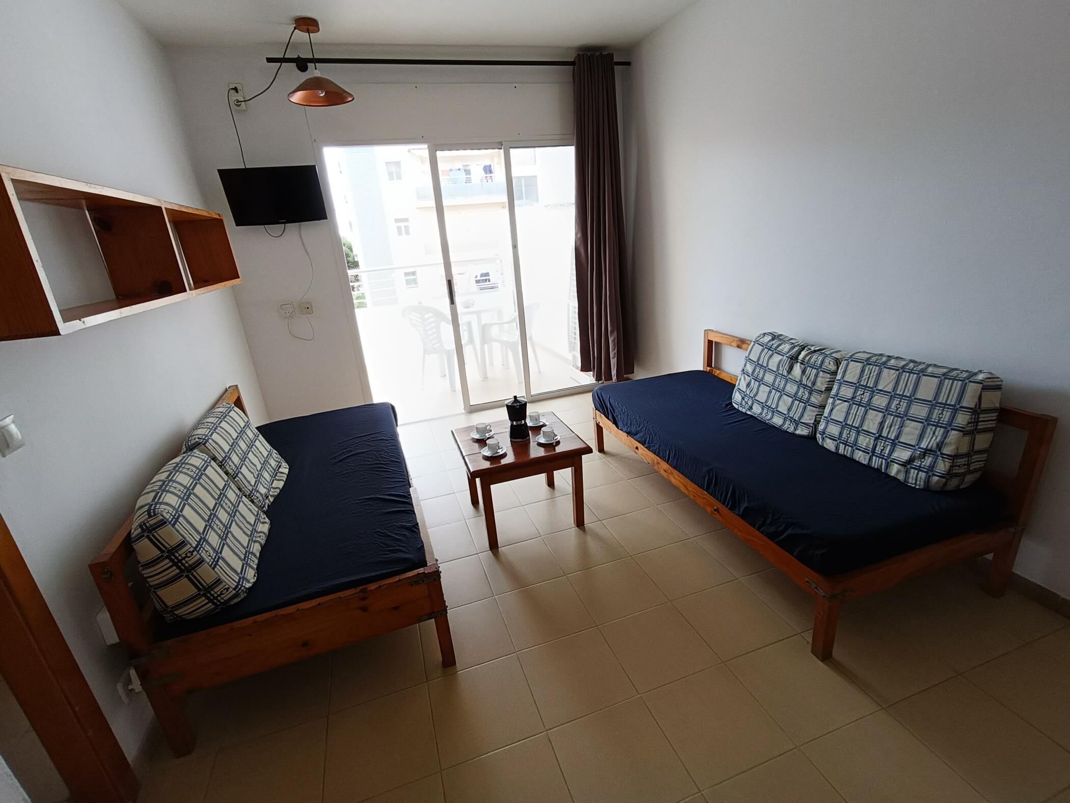 A small, well-lit room with two wooden beds, a small table with a coffee maker, a TV mounted on the wall, and a sliding glass door leading to an outdoor area.