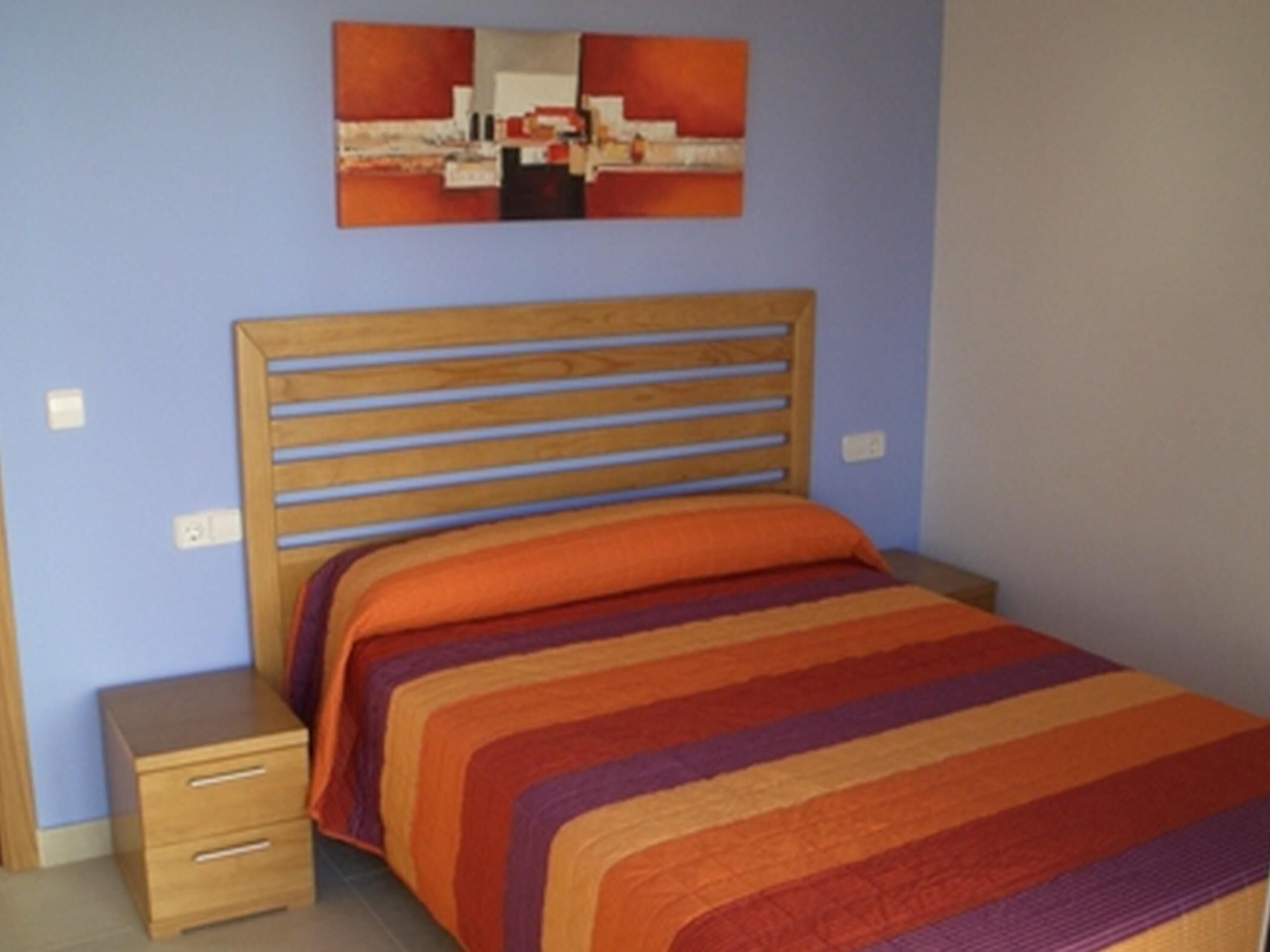 A bedroom with a wooden bed frame, a colorful striped bedspread, a nightstand with drawers, and a wall-mounted abstract painting.