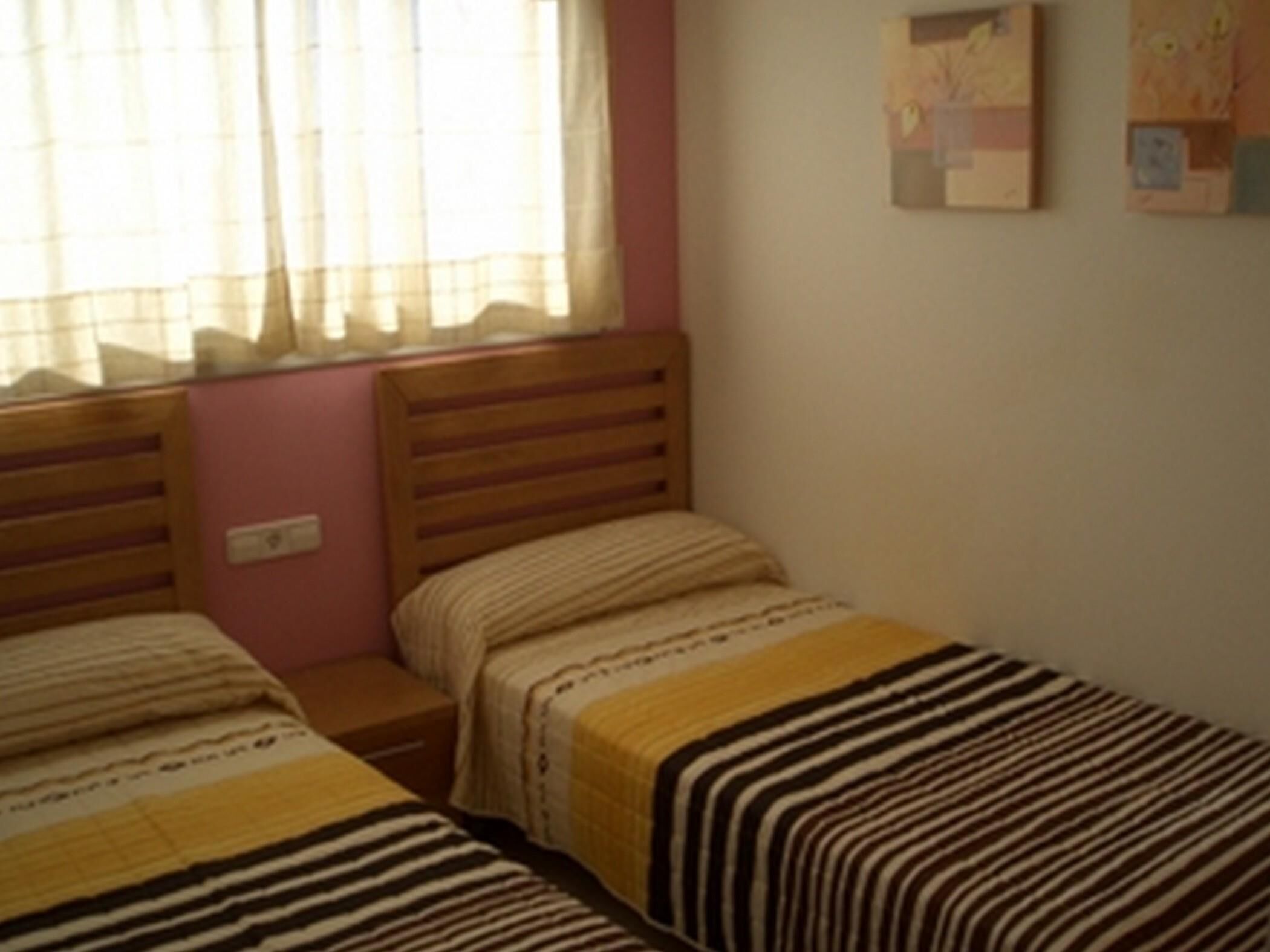 Un dormitorio con dos camas individuales, cabecera de madera, mesita de noche y ventana con cortinas traslúcidas.