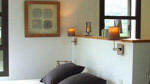 Ein Schlafzimmer mit einem Bett, zwei Nachttischlampen, einem gerahmten Kunstwerk über dem Bett und einem Fenster mit Blick.