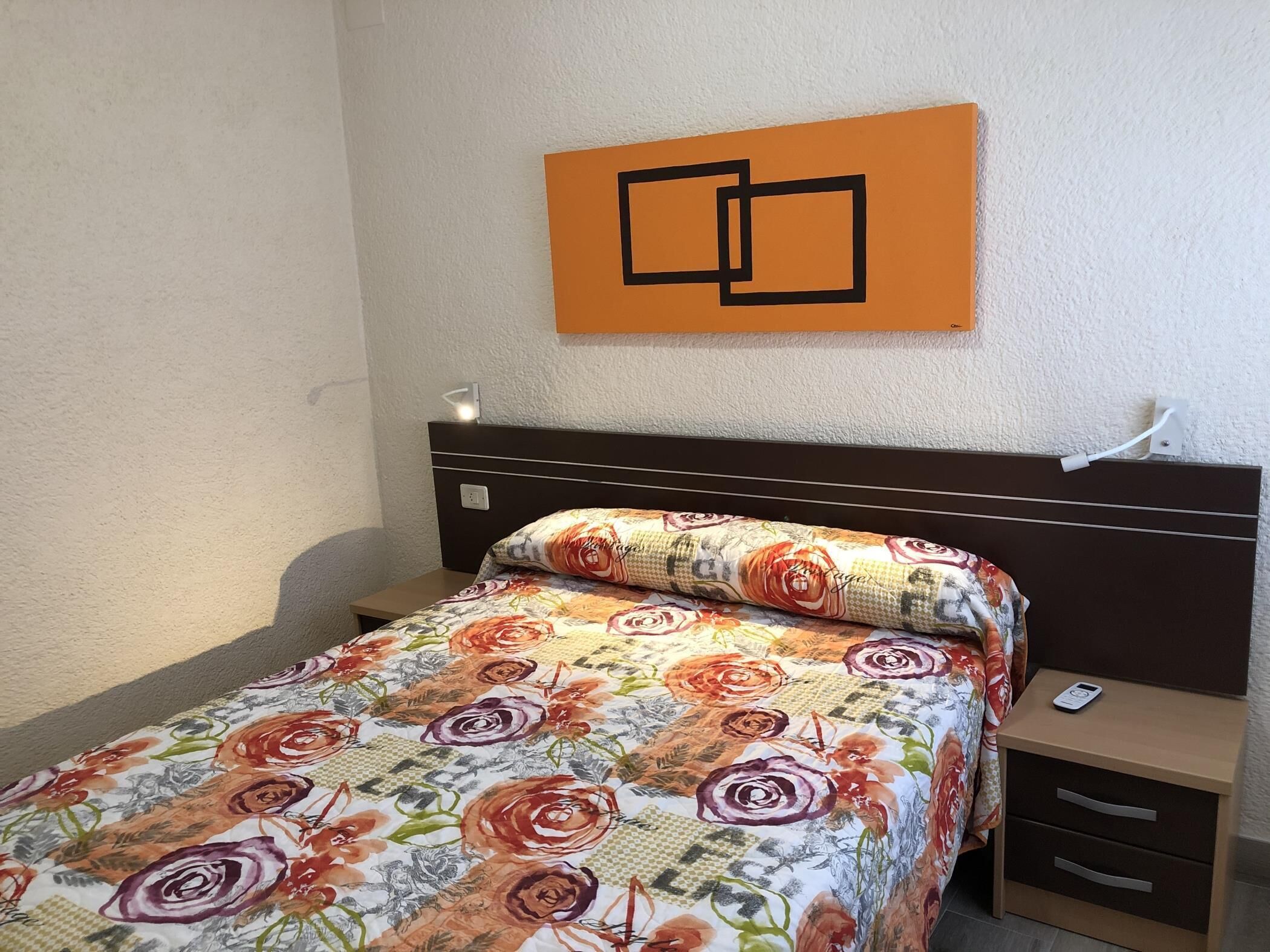 Une chambre d’hôtel comprenant un lit, une table de chevet, un tiroir, une lampe fixée au mur et une œuvre d’art décorative.