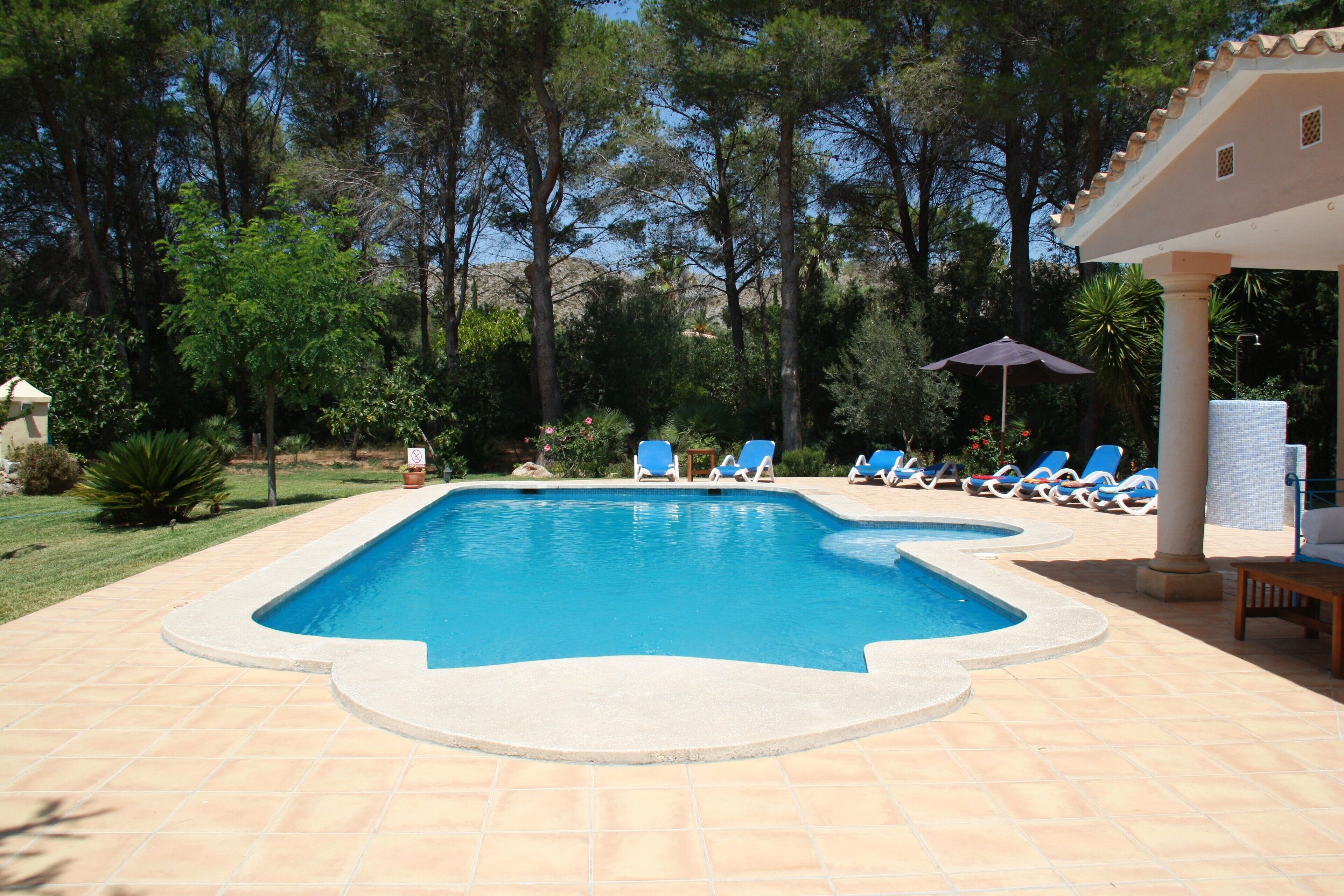 Une piscine rectangulaire, bordée d’une terrasse carrelée, entourée de chaises longues et agrémentée d’une pergola avec un parasol.