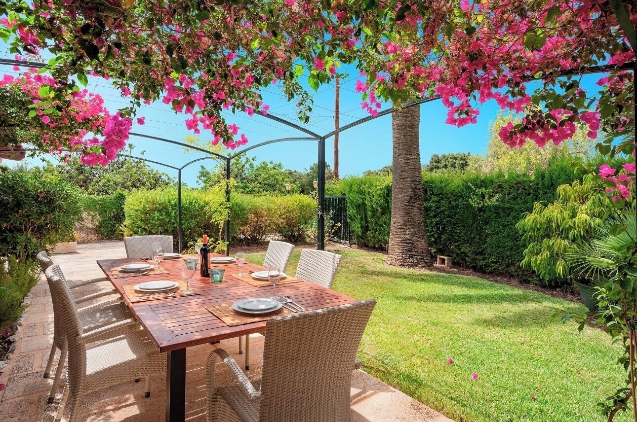 Un'area pranzo all'aperto con tavolo in legno e sedie in vimini, circondata da piante rigogliose e fiori.
