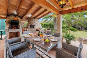An outdoor patio with a stone fireplace, wicker furniture, and a table set for breakfast. - Beautiful very quiet villa with pool, large terraces and a large garden. (Pollença)