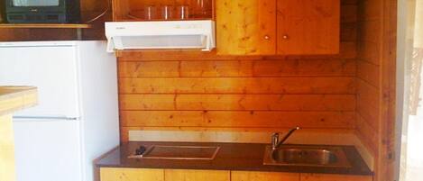 Una cocina compacta con armarios de madera, un refrigerador blanco, un lavamanos y un televisor pequeño encima del refrigerador.