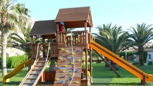 Une structure de jeux en bois, dotée d’un toboggan et d’un mur d’escalade, entourée de pelouse et de palmiers.