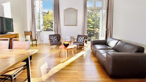 Ein Wohnzimmer mit einer Ledersofa, Holzboden, einem Fernseher auf einem Holztisch, zwei Stühlen mit gemusterten Kissen und großen Fenstern mit Vorhängen.