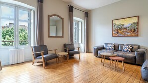 Ein geräumiges Wohnzimmer mit Holzboden, einer grauen Couch mit gemusterten Kissen, einem Couchtisch aus Holz und zwei Sesseln. Ein großer Spiegel hängt an der Wand, und über der Couch ist ein Gemälde zu sehen.
