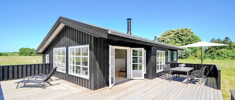 A modern wooden cabin with a deck, a patio area with a table and chairs, and a large open door leading to an interior space.